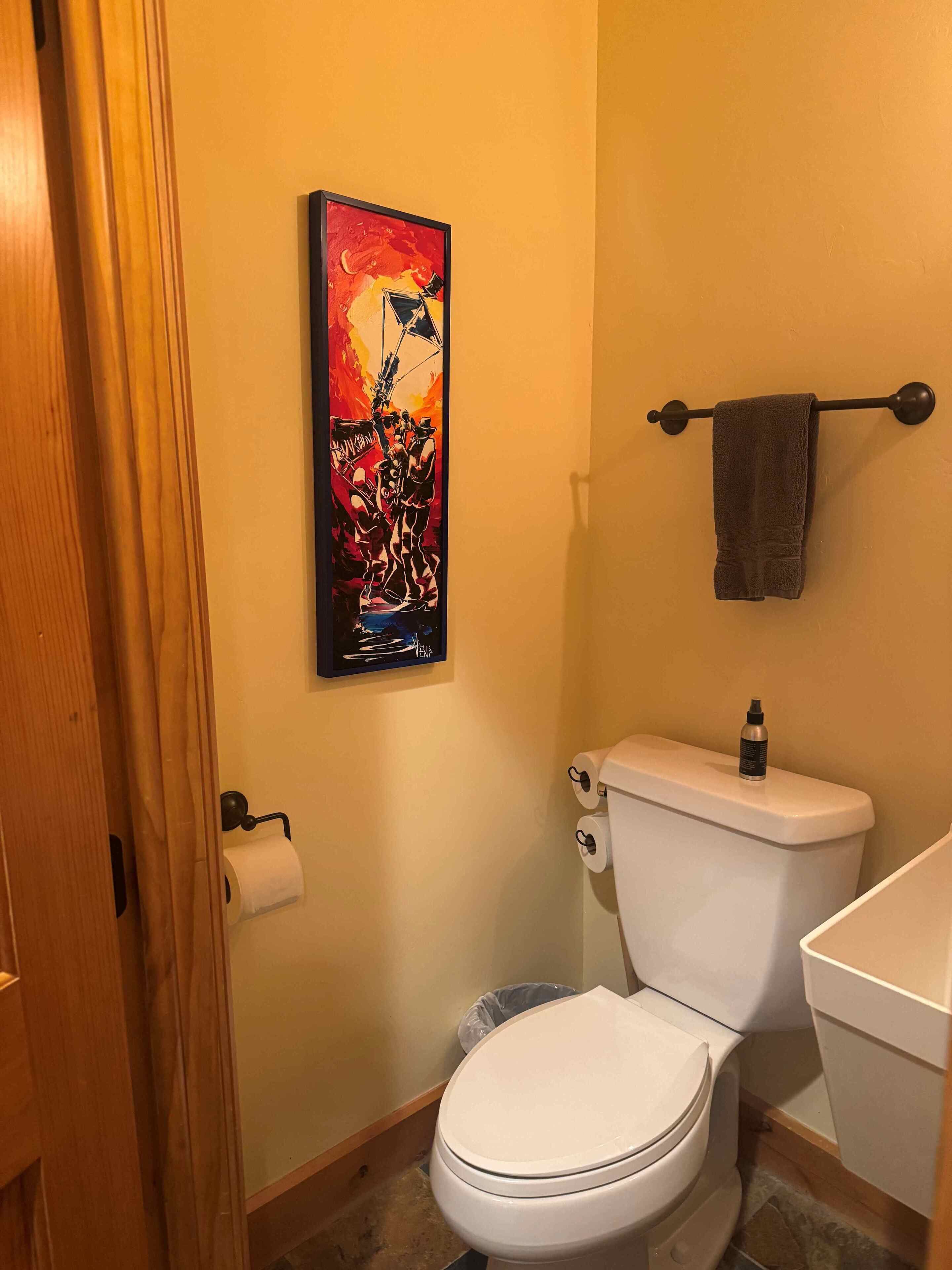 A bathroom with a toilet, a towel rack, a painting, and a roll of toilet paper.