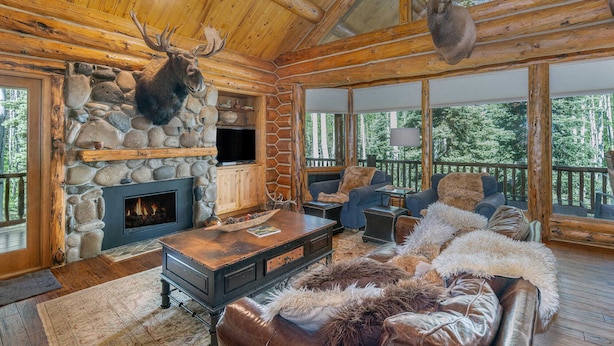 L’intérieur d’une cabane en rondins, avec une cheminée en pierre, un téléviseur et un coin salon confortable agrémenté d’un plaid en fourrure.
