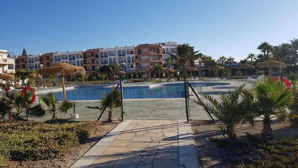 Zwembadgedeelte van een resort, met een helderblauw zwembad, omringd door palmbomen en een hek.