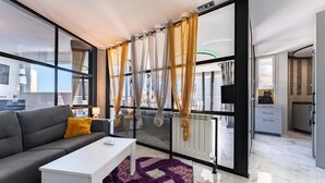 Un salon moderne avec un canapé gris, une table basse blanche et un tapis violet. De grandes fenêtres avec des rideaux légers offrent une vue sur la ville.