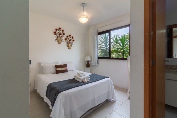 A hotel room with a bed, a window with a view of greenery, a wall-mounted light fixture, and a small table with a lamp.