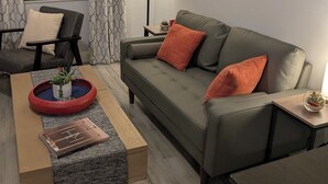 Een moderne woonkamer met een grijze bank, een houten salontafel, een bijzettafeltje met een lamp en een raam met patroon gordijnen.