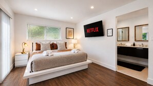 Ein modernes Schlafzimmer mit Bett, Nachttischen, einem Fernseher und einem Badezimmer mit Waschbecken und Spiegel.