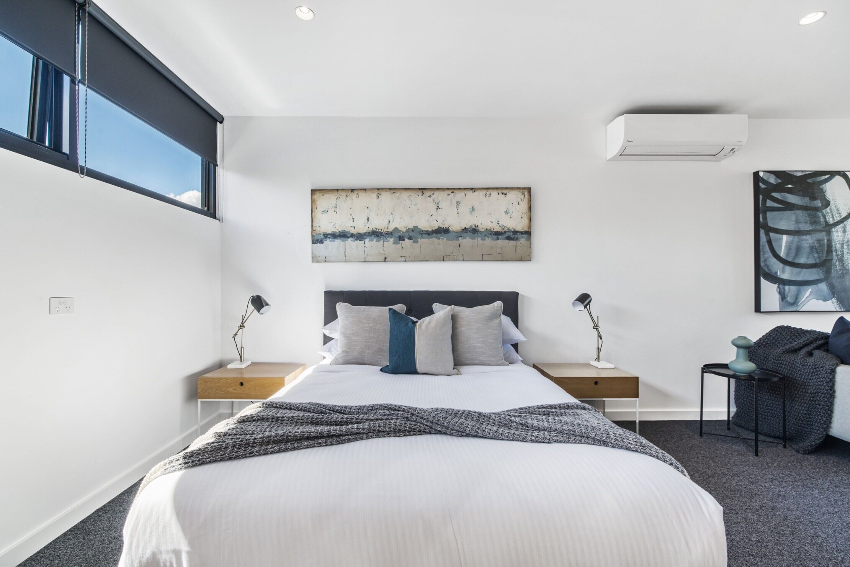 A modern bedroom with a large bed, a headboard, bedside tables with lamps, a wall-mounted air conditioner, and a window with blinds.