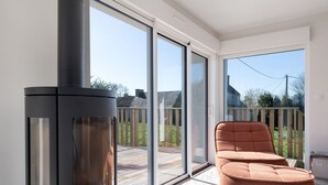 Una habitación moderna con un gran ventanal corredero que da a un balcón, una estufa de leña de madera negra y un sillón tapizado marrón.