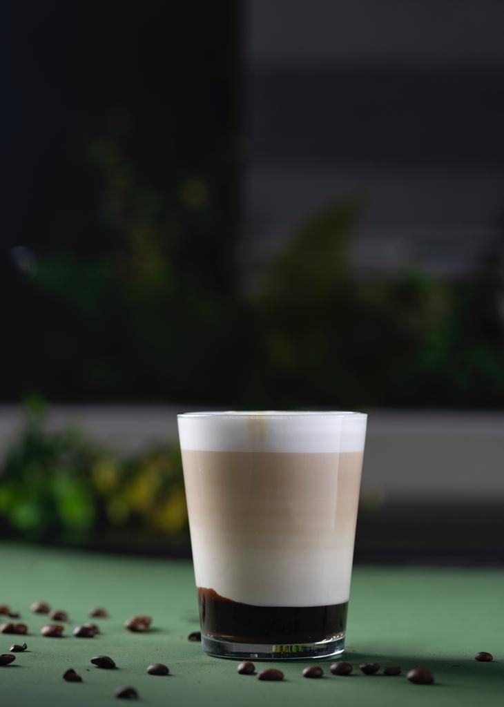 A glass of layered beverage with coffee and milk, placed on a green surface with scattered coffee beans.
