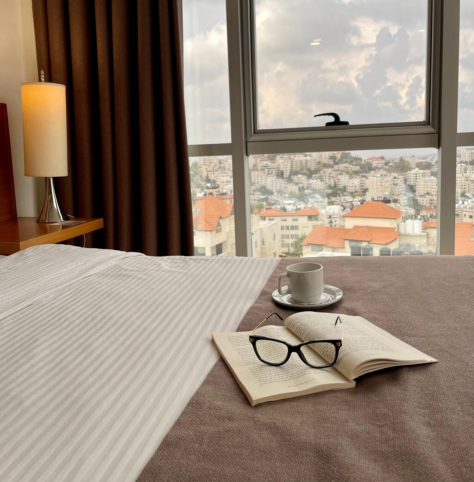 Une chambre d’hôtel avec un lit, une lampe de chevet, une tasse de café et un livre ouvert, sur lequel reposent des lunettes.