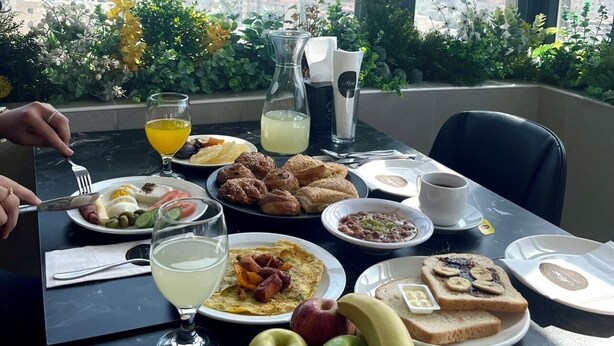 A breakfast table with various dishes, a glass of orange juice, and a view of the city through large windows. - Caesar Hotel (Ramallah)