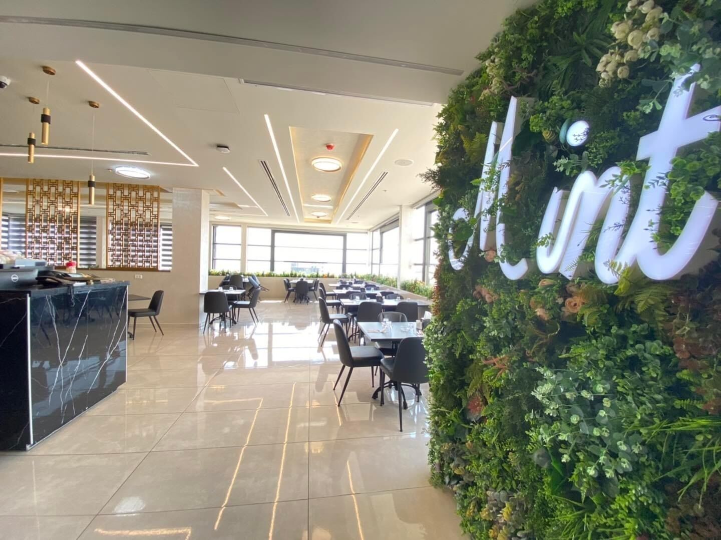 A spacious indoor area with a reception desk, multiple tables, and chairs arranged for dining or meetings. A large wall with greenery and the word "mint" is visible.
