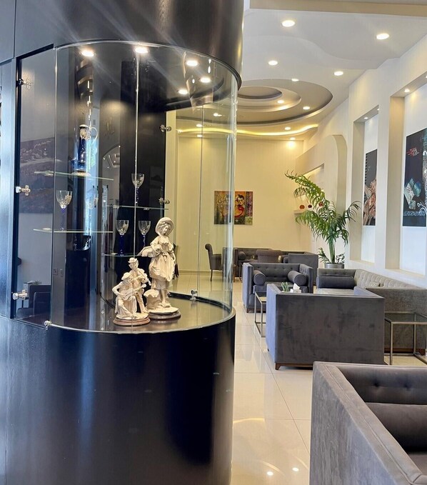 Un lobby moderno con un expositor de vidrio central que contiene objetos decorativos, una zona de estar con sofás y una planta grande.