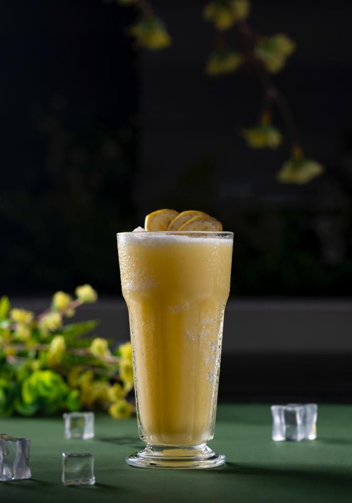 A tall glass with a yellow beverage, ice cubes, and a lemon slice on a green surface.