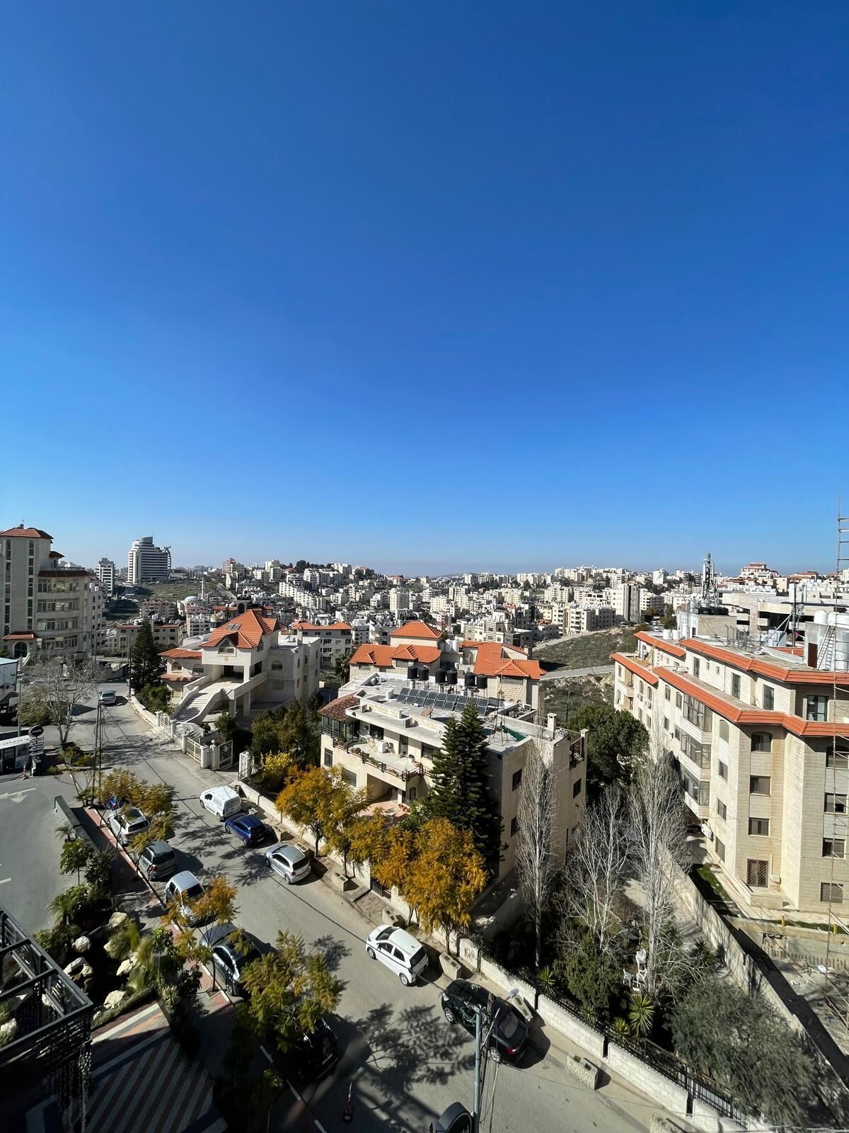 A cityscape with residential buildings, a clear blue sky, and a road with parked cars.