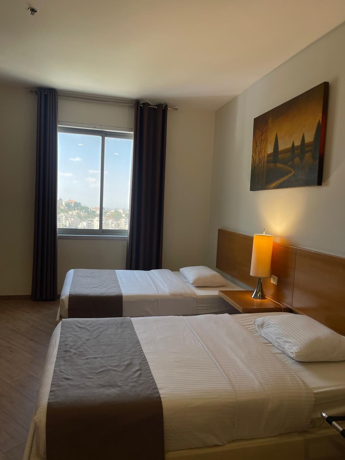Habitación de hotel con dos camas, un ventanal con cortinas, un cuadro en la pared y una lámpara de noche.