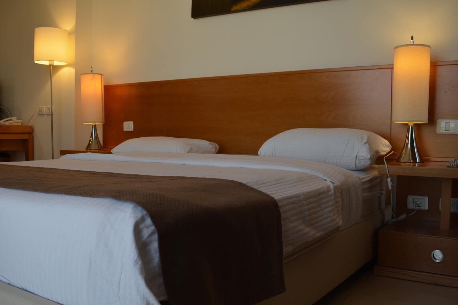 A hotel room with a large bed, two bedside lamps, a wooden headboard, and a nightstand with a phone and a lamp.