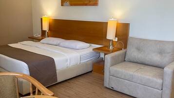A hotel room with a bed, two bedside tables with lamps, a sofa, a round wooden table with a tissue box and glass, and a painting of elephants on the wall.