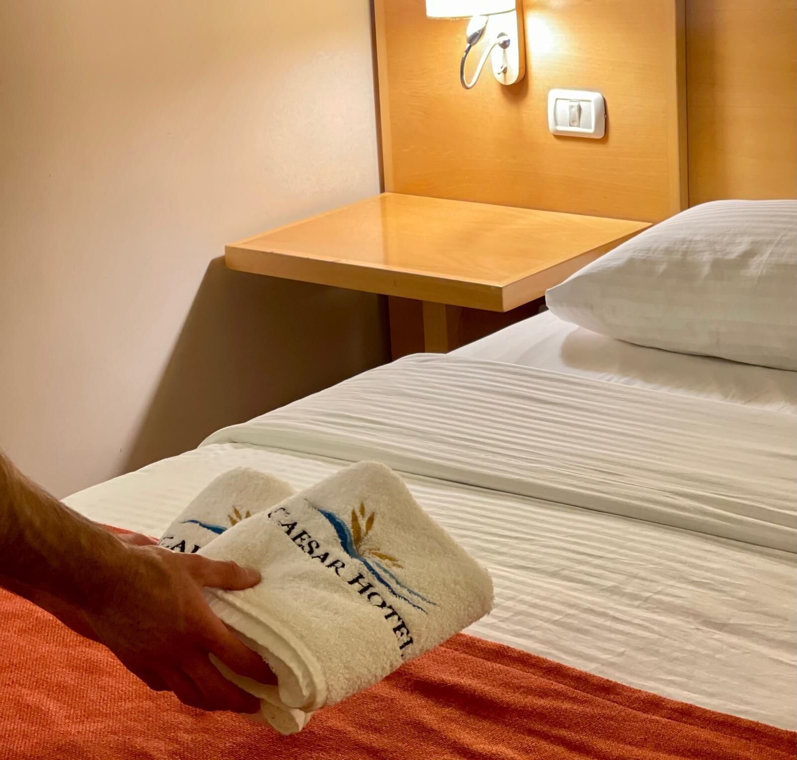 A hotel room with a neatly made bed, a wooden nightstand with a lamp, and a hand holding a towel with 'CAESAR HOTEL' embroidered on it.