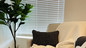 A beige sofa with a white throw blanket and a black pillow, positioned in front of a window with blinds. - The Emerald Escape 2BR/2BA King Suite (Hermiston)