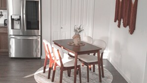 A dining area with a wooden table and chairs, a white rug, a refrigerator, and a white door with a decorative wall hanging. - The Emerald Escape 2BR/2BA King Suite (Hermiston)