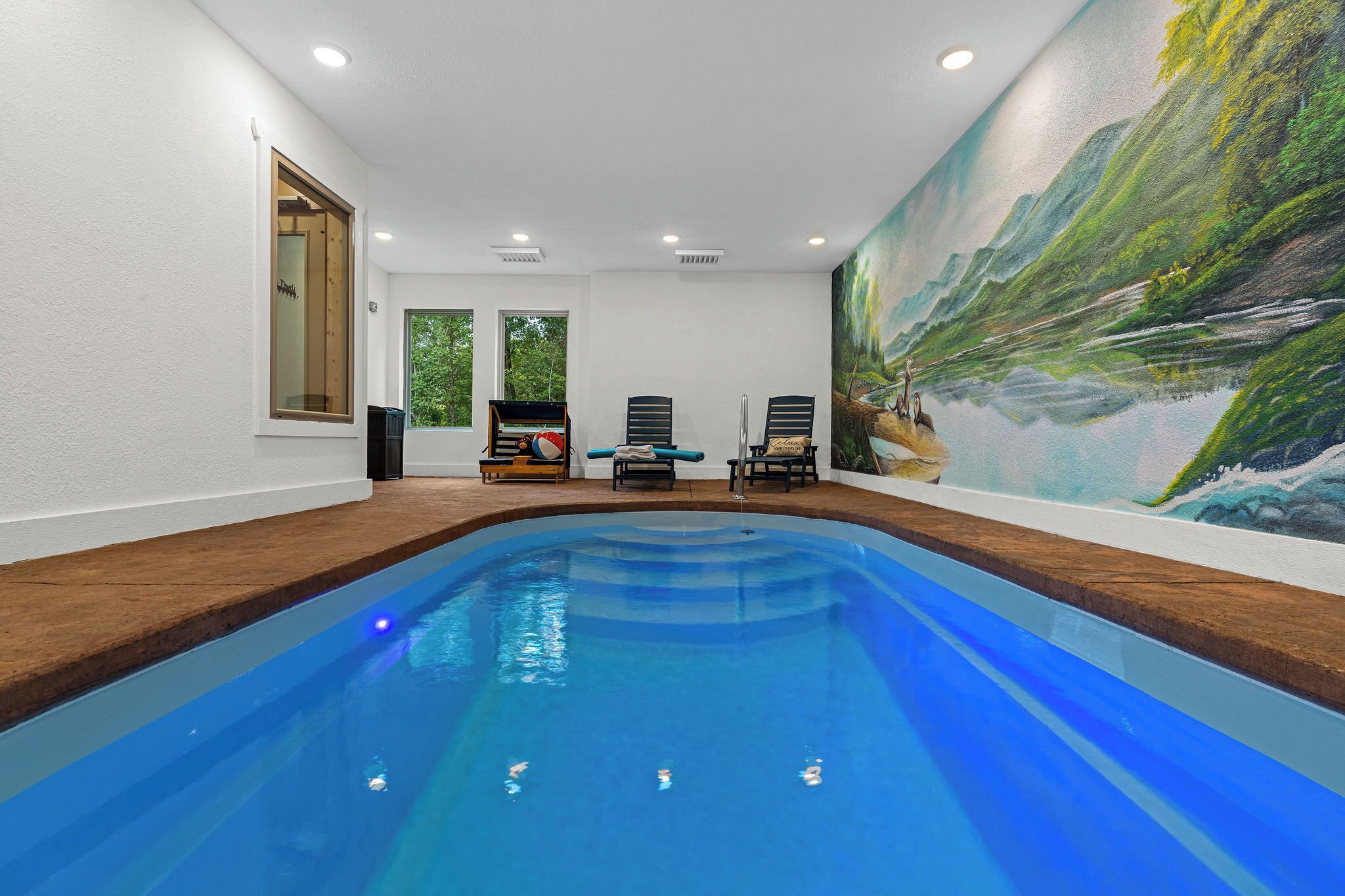 Una piscina interior con un mural, un muelle de madera y dos sillones de descanso.