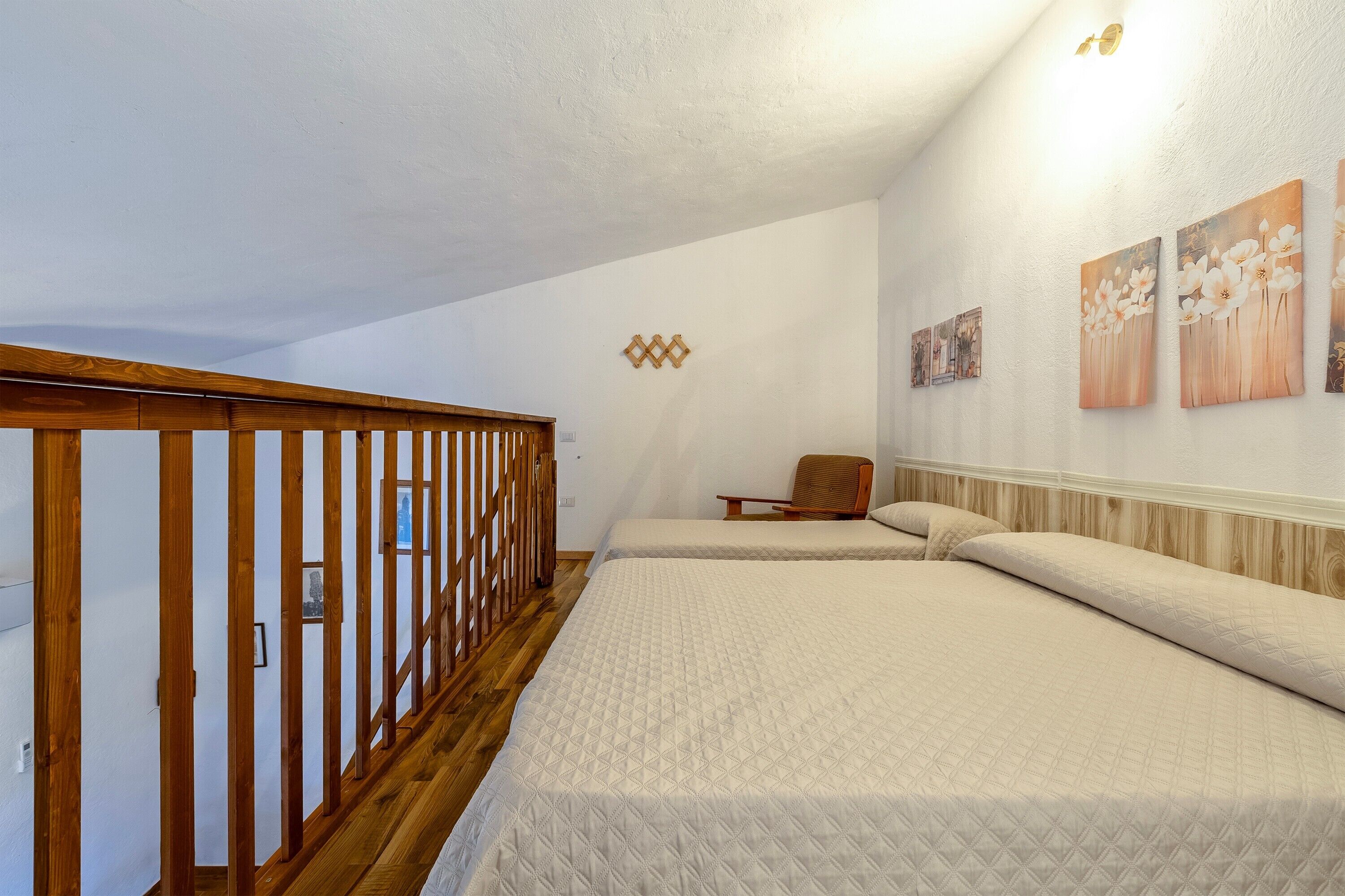 A bedroom with two beds, a wooden railing, a chair, and wall art.