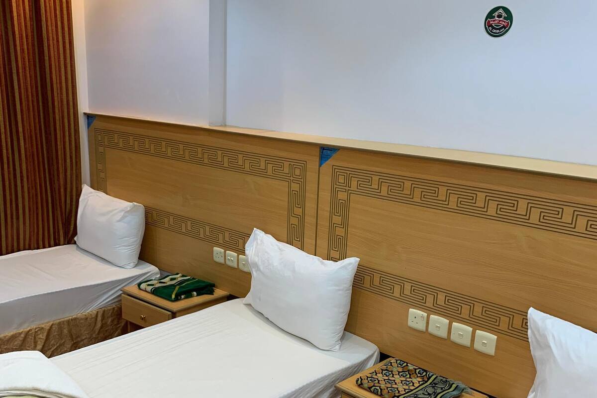 Une chambre d’hôtel avec deux lits simples, chacun doté d’un oreiller blanc et d’une couverture verte. On y trouve une tête de lit en bois ornée d’un motif décoratif, ainsi qu’une petite table de chevet recouverte d’un tissu à motifs.