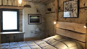 Une pièce rustique avec un plafond en bois, un lit recouvert d’une courtepointe, une fenêtre et deux tableaux encadrés accrochés au mur.