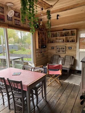 A wooden interior with a table and chairs, a bench, hanging plants, and shelves with various items.