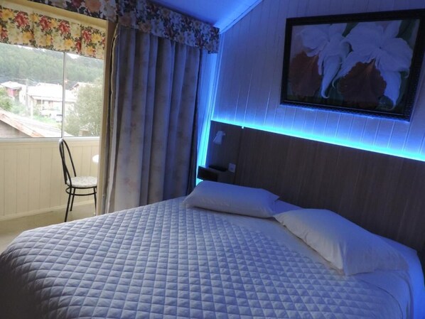 Une chambre à coucher comprenant un lit, une table de chevet, une chaise, une fenêtre avec des rideaux et un tableau accroché au mur.