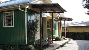 A green building with a covered porch, a small table and chair, and a paved walkway. - Cabanas Costaneira  (Cambará do Sul)