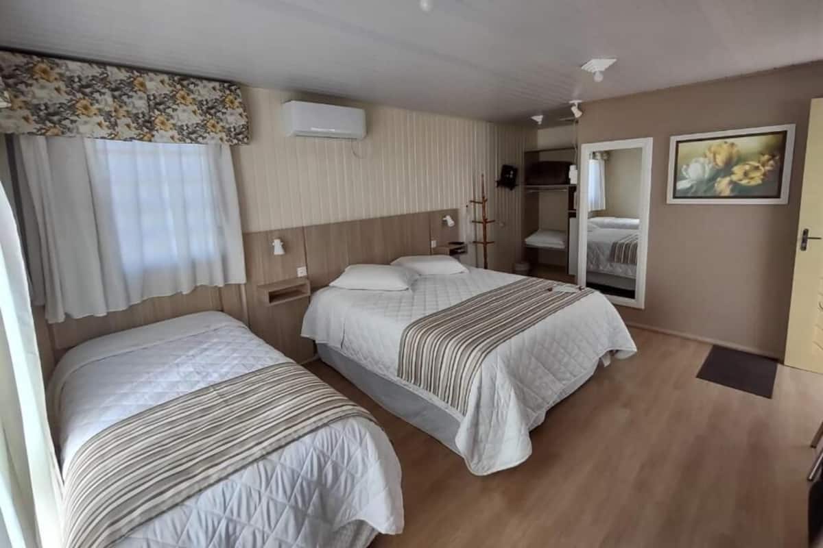 A hotel room with two beds, a wooden floor, a window with curtains, an air conditioning unit, a painting, and a door.