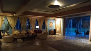 A spacious living area with a large carpet, sofas, and a coffee table. There is a large window with curtains, a painting on the wall, and a view of the outside. - Palais Oussoud Marrakech (Marrakech)