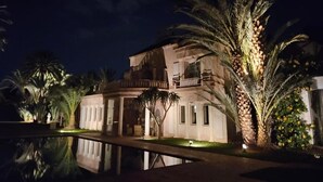 A multi-story building with a pool in front, surrounded by palm trees and well-lit at night. - Palais Oussoud Palmeraie (Marrakech)