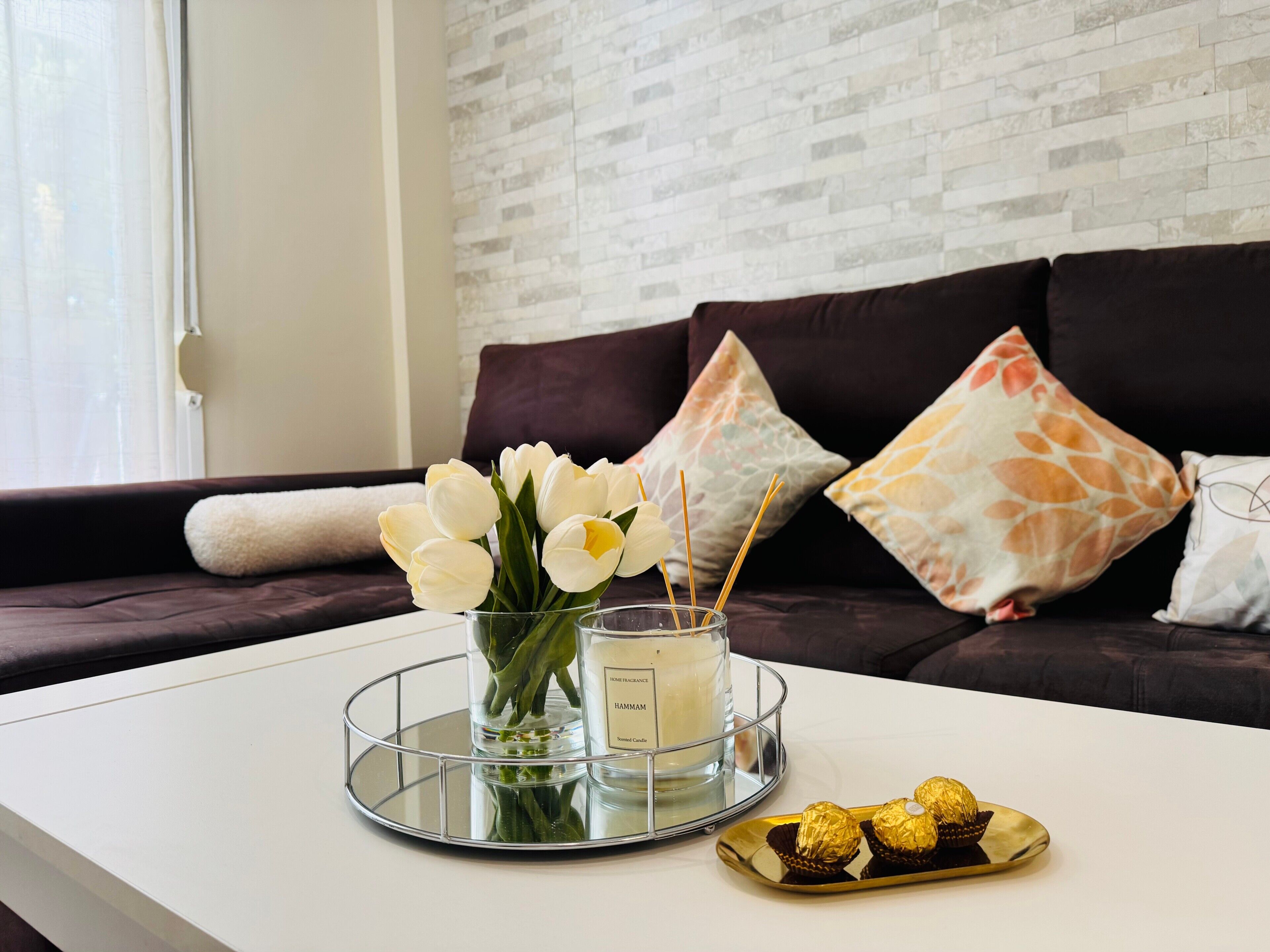 A modern living room with a white coffee table, a glass candle holder with white flowers, and a tray with chocolates.