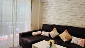 Una sala de estar con un sofá marrón oscuro, cojines blancos, una mesa de centro de vidrio con un florero y una pared con diseño de ladrillo.