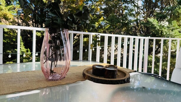 Una mesa de cristal con un florero de hojas verdes, un posavasos de madera y una guirnalda de luces en un balcón.