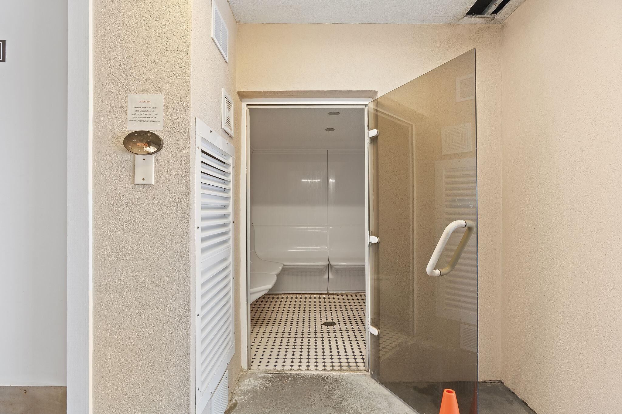 Corredor estreito que dá para uma área de chuveiro, com porta de vidro, sistema de ventilação e piso revestido de azulejos.