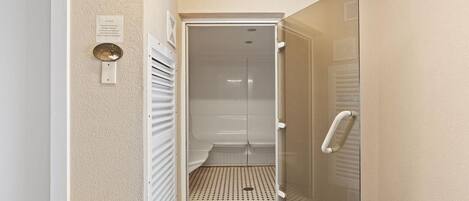 Corredor estreito que dá para uma área de chuveiro, com porta de vidro, sistema de ventilação e piso revestido de azulejos.