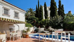 Un'area piscina con un edificio bianco, posti a sedere all'aperto e alte cipressi.