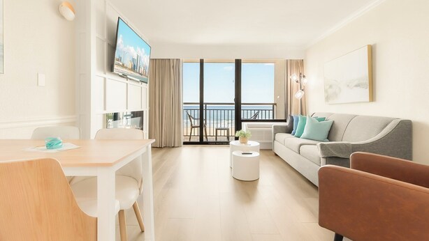 A modern living room with a dining area, a sofa, a coffee table, and a TV. There is a balcony with a view of the sea. - The Caribbean Resort and Villas #327 (Myrtle Beach)