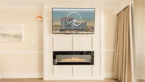 A living room with a fireplace, a mounted TV displaying 'WAVECREST BEACH VACATIONS', and a beach view. - The Caribbean Resort and Villas #327 (Myrtle Beach)