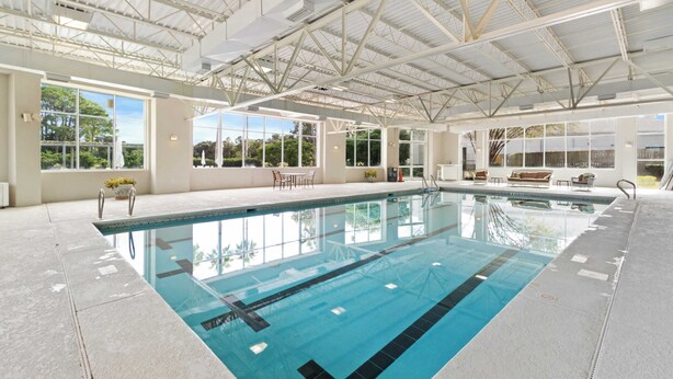Une piscine intérieure avec des parois en verre transparent, un plafond haut avec des poutres métalliques et une vue sur la verdure environnante.