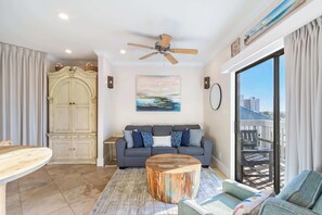 A living room with a grey sofa, a round wooden coffee table, a ceiling fan, and a large window with a view of buildings. - 1 bed Beach, Pools, Waterfront, Boating | Palmaway (Orange Beach)