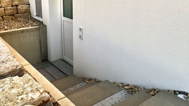 Eine schmale Treppe führt zu einer Tür, eine Steinwand und eine weiße Wand mit einer Leuchte.