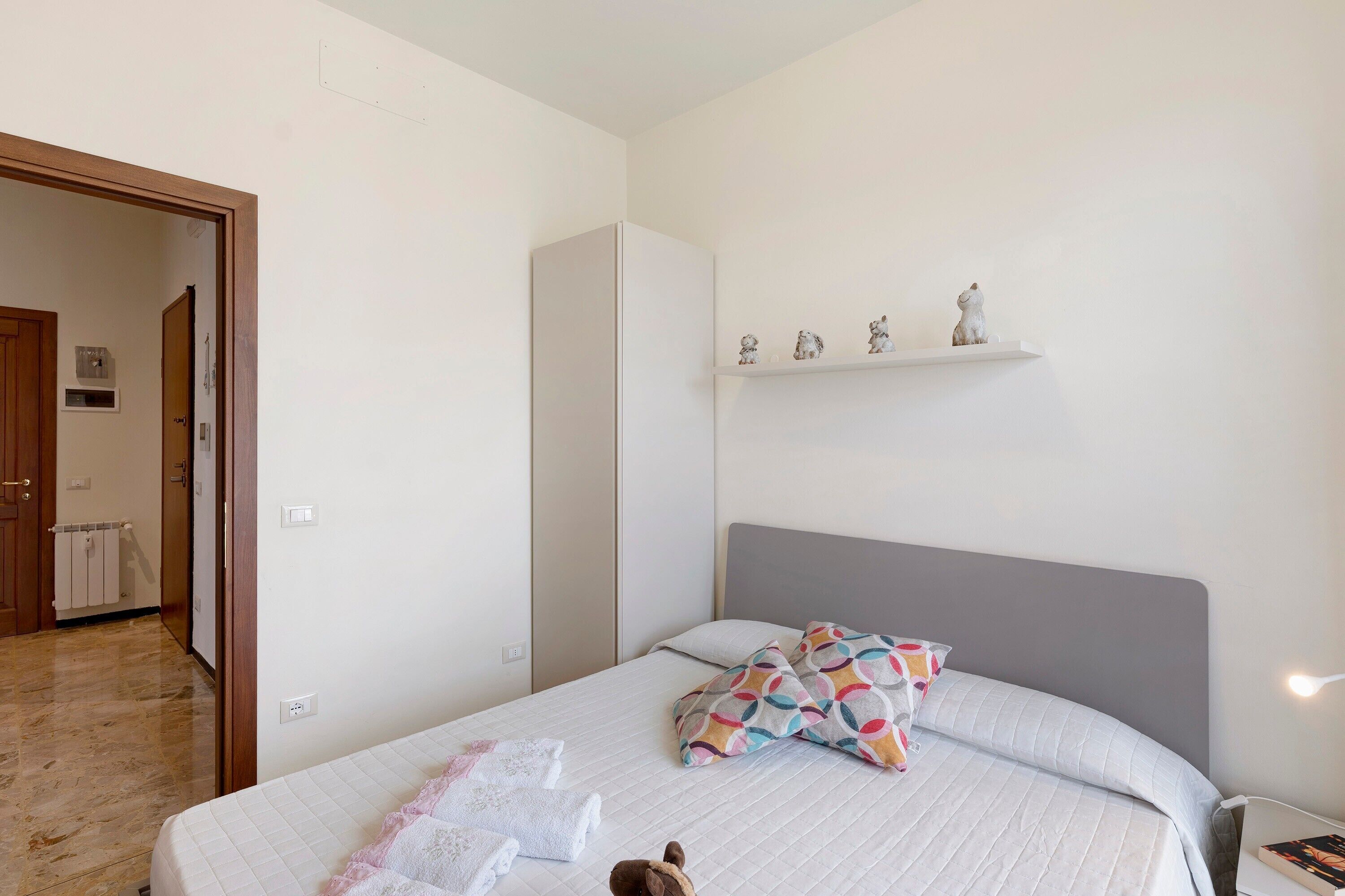 Un dormitorio con cama, mesita de noche, repisas con objetos decorativos y una puerta que da a otra habitación.