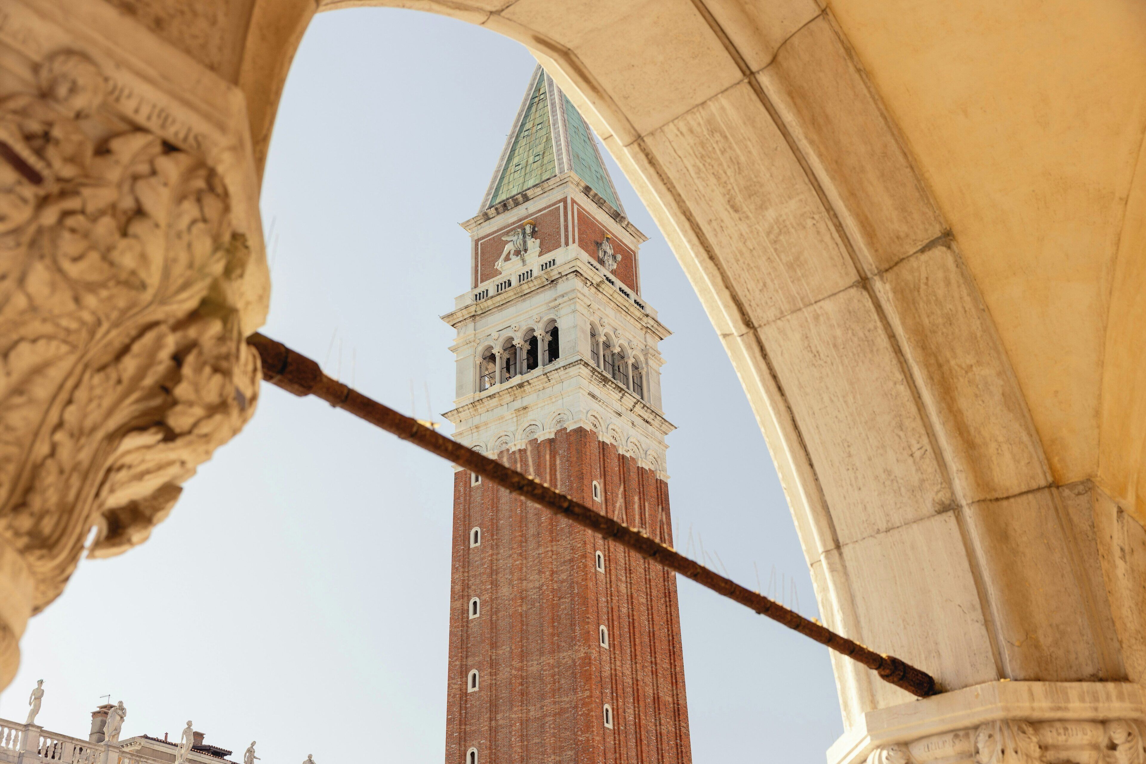 A tall brick tower with a clock and a green roof, viewed through an ornate archway.