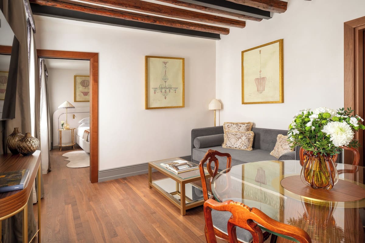 Una sala de estar moderna con una mesa de centro de vidrio, un sofá gris y una mesa de comedor de madera con sillas. Hay obras de arte enmarcadas en las paredes y un jarrón con flores sobre la mesa de comedor.