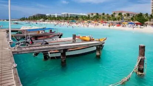 Vista de la playa con un muelle, barcos y cielo azul despejado.