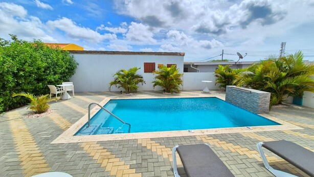 A rectangular swimming pool with a metal ladder, surrounded by a tiled patio with a herringbone pattern. There are two lounge chairs and a small table with a chair. - Charming 1-bedroom apartment in perfect Oranjestad with outside Pool (Oranjestad)