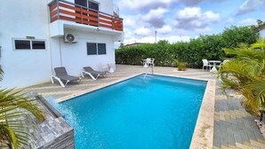 Una piscina con sillones de descanso y una mesita, rodeada de vegetación y un edificio blanco con balcón.