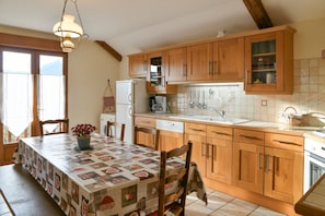 Une cuisine avec des meubles de rangement en bois, une table à manger recouverte d’une nappe à motifs et un luminaire suspendu.